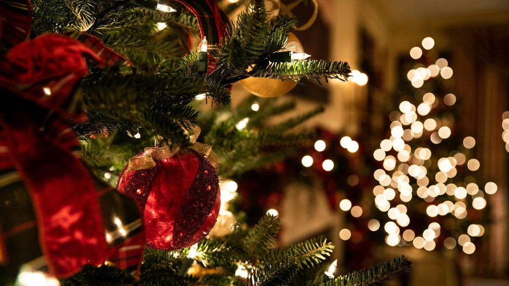 Close-up of a decorated Christmas tree with red ornaments and ribbons, with another lit tree blurred in the background.