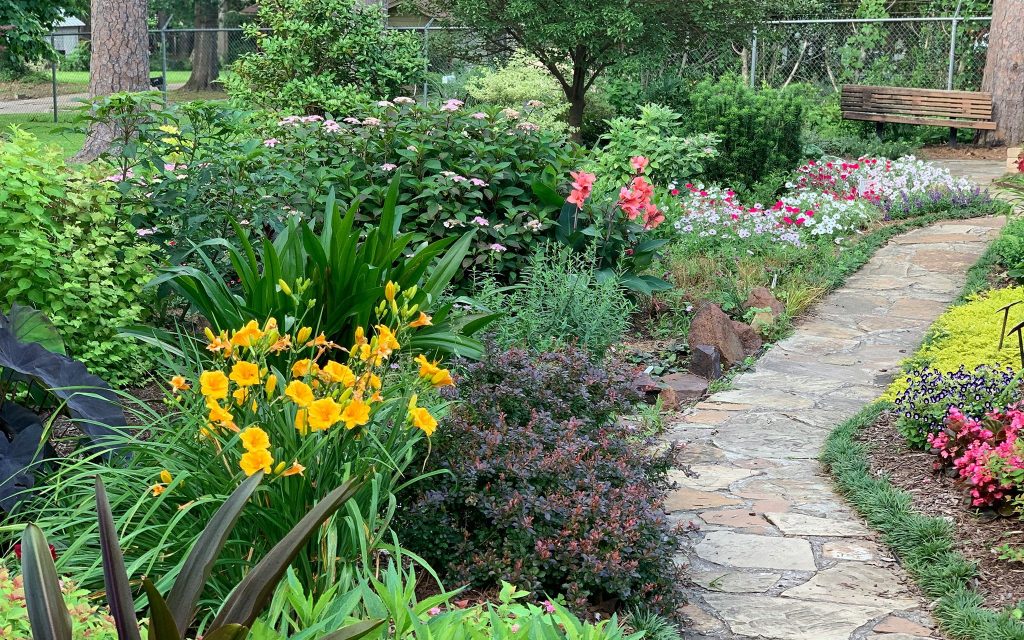 Stone walkway curves through a lush garden with yellow, pink, and red flowers, green shrubs, and a wooden bench in the background near a fence.